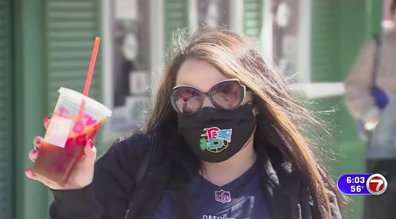 A woman holding a Dunkin' iced coffee and wearing a Patriots jersey and New England sports facemask