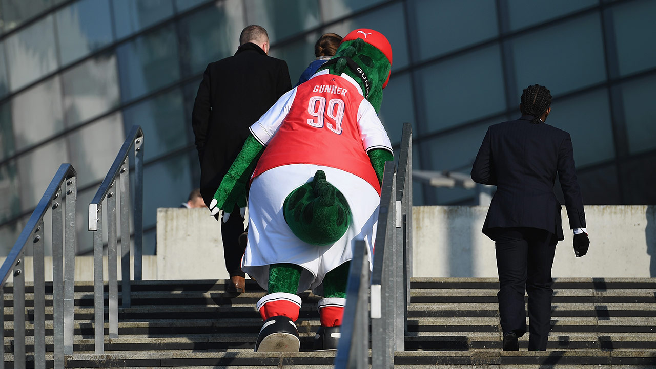 Gunnersaurus, the Arsenal mascot, which is a big green dinosaur in a red Arsenal jersey, walks up the steps escorted by some people in suits.