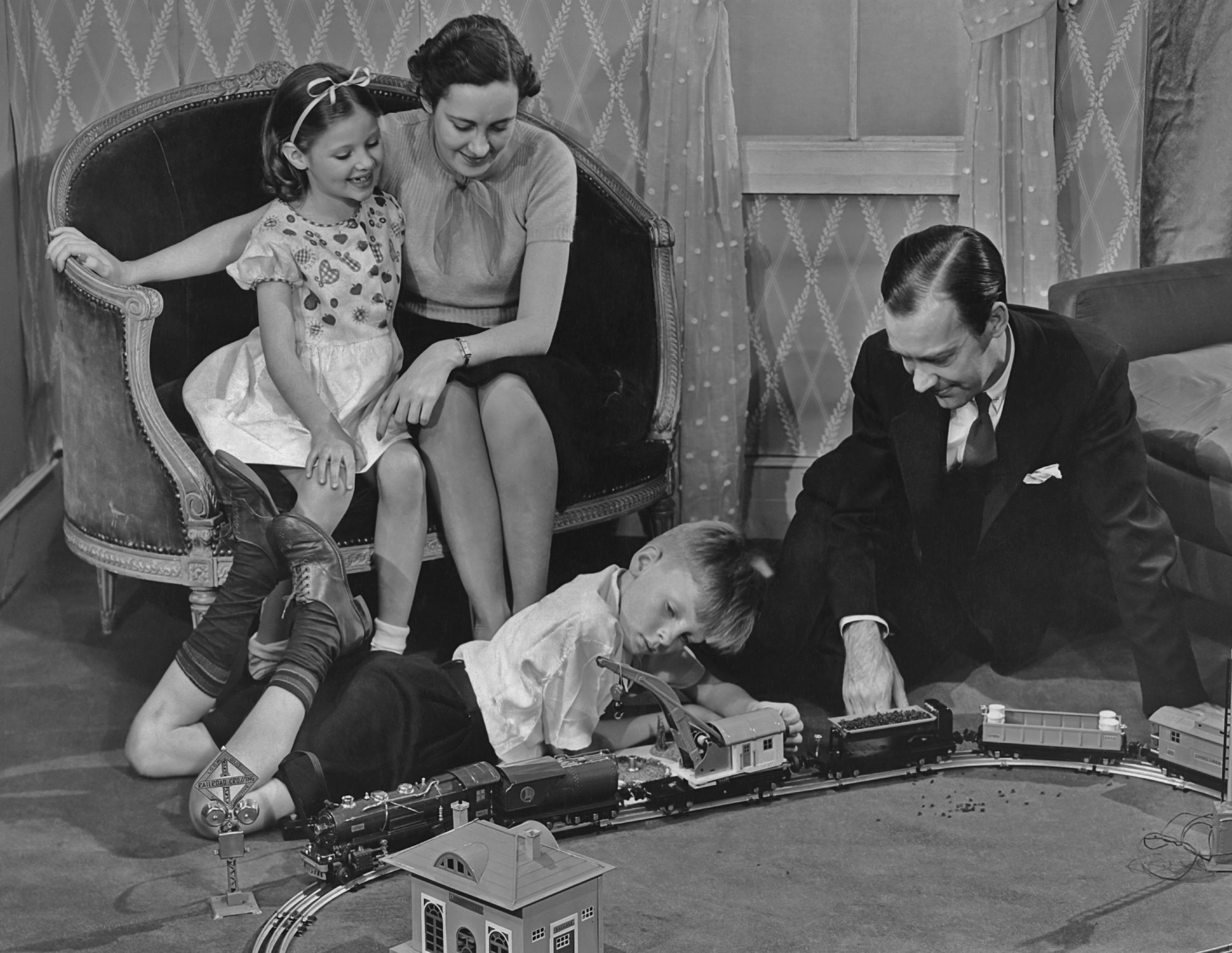 Father and son play with a toy train set as mother and daughter look on circa 1940's.
