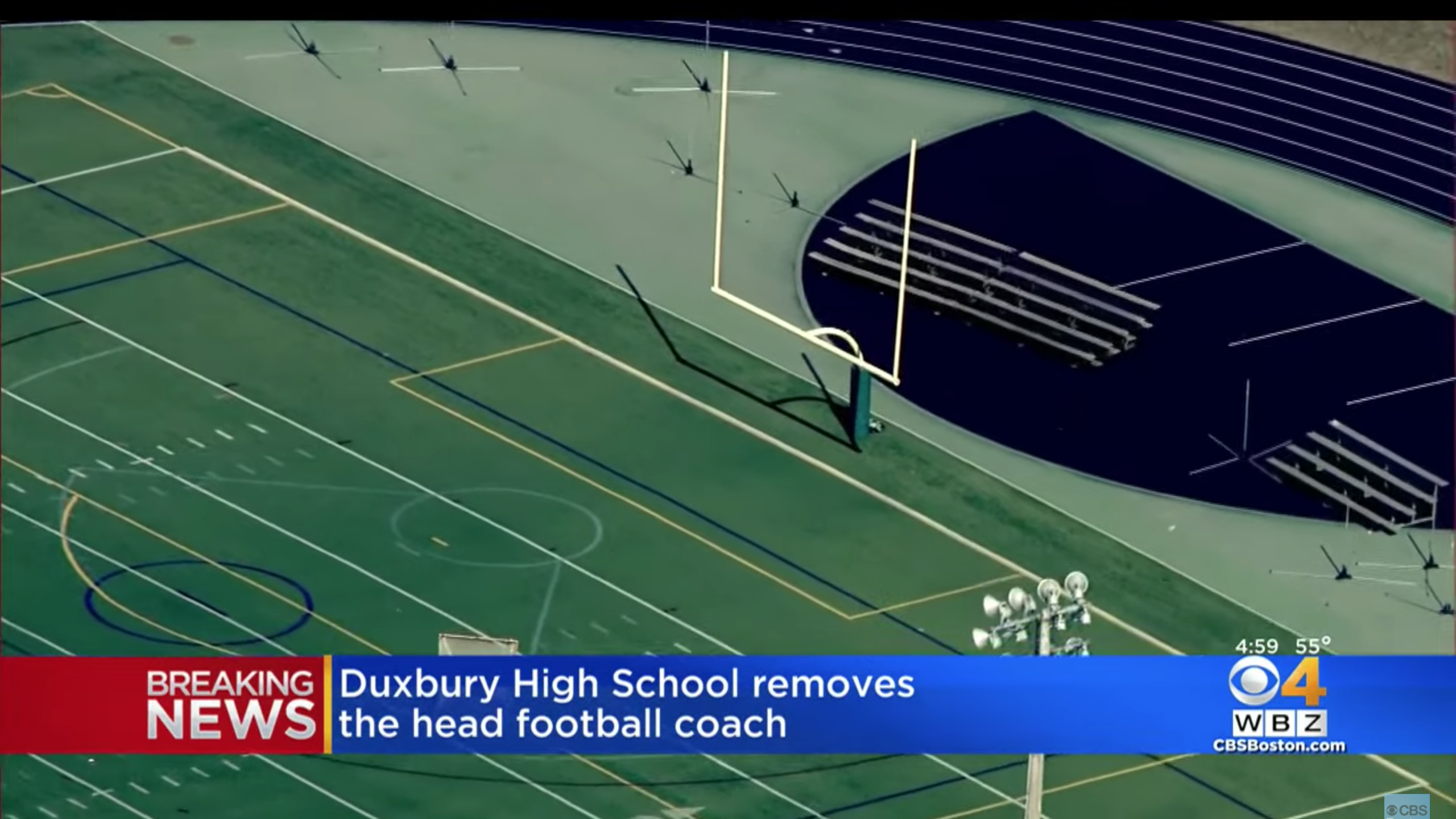 The football field at Duxbury High School. You can see the field, a track around it, and the goal posts. There's also a TV graphic that says "Breaking News: Duxbury High School removes the head football coach."