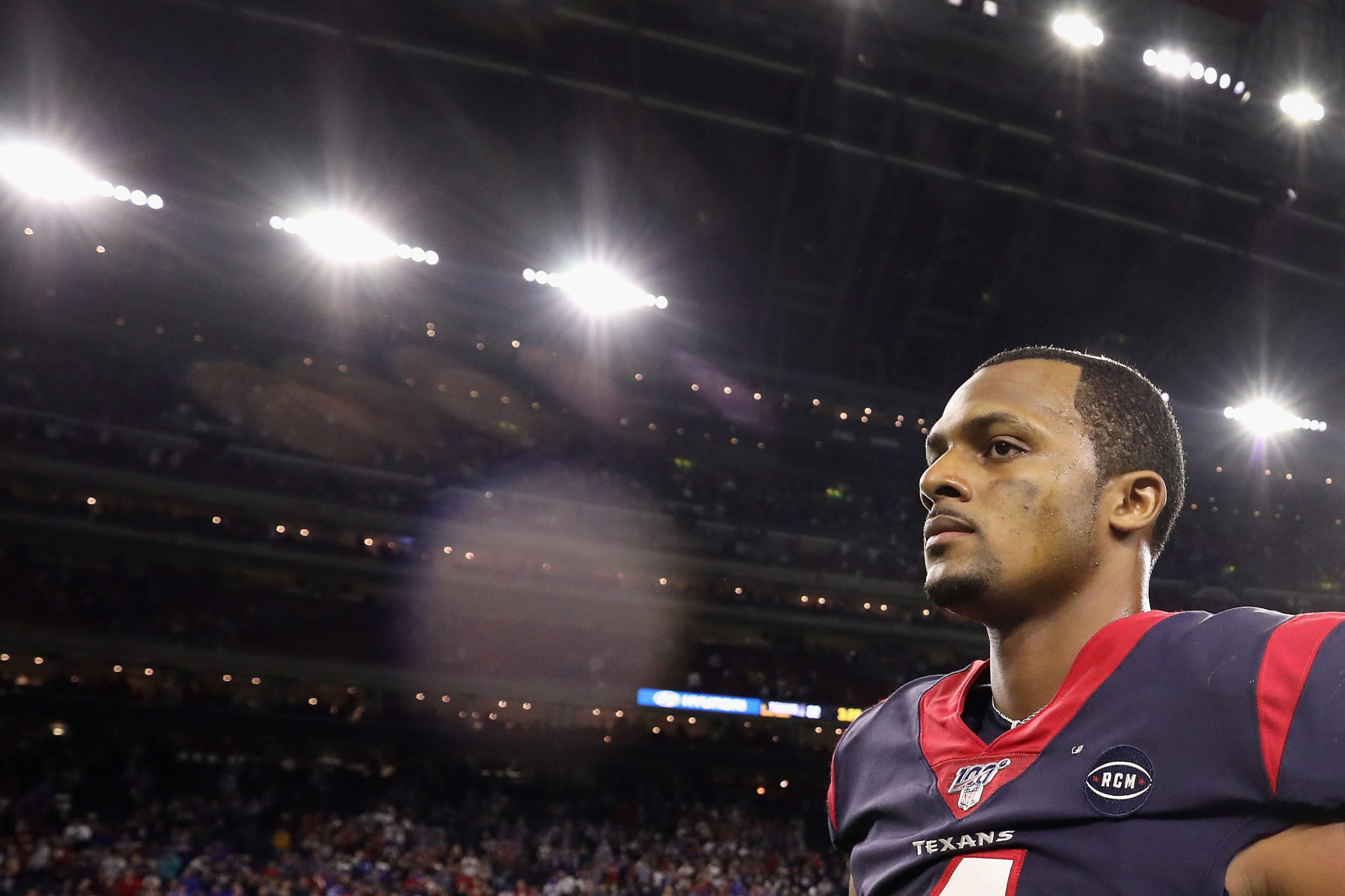 Quarterback Deshaun Watson #4 of the Houston Texans walks off the field following the AFC Wild Card Playoff game against the Buffalo Bills at NRG Stadium on January 04, 2020 in Houston, Texas. Watson is dressed in his uniform. In the background are stadium lights and fans.