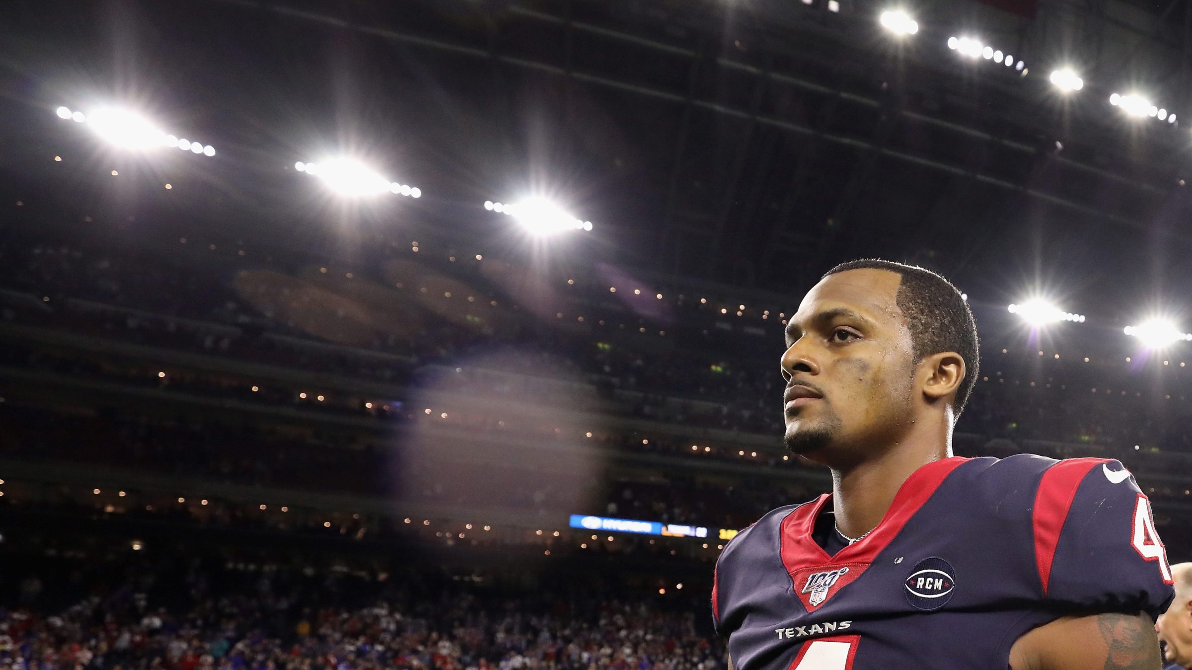 Quarterback Deshaun Watson #4 of the Houston Texans walks off the field following the AFC Wild Card Playoff game against the Buffalo Bills at NRG Stadium on January 04, 2020 in Houston, Texas. Watson is dressed in his uniform. In the background are stadium lights and fans.