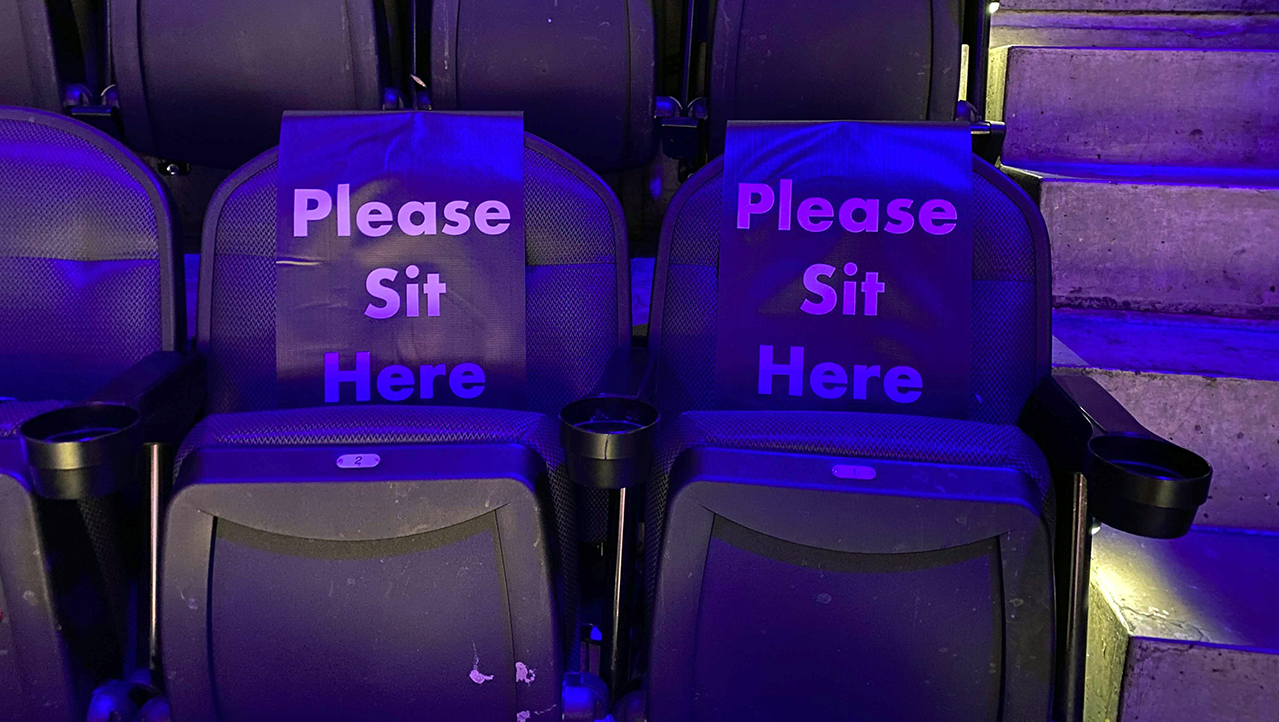 Two seats in an arena at an NBA game. They are black. There are steps next to them. Both seats have a cover on them that says 'PLEASE SIT HERE'