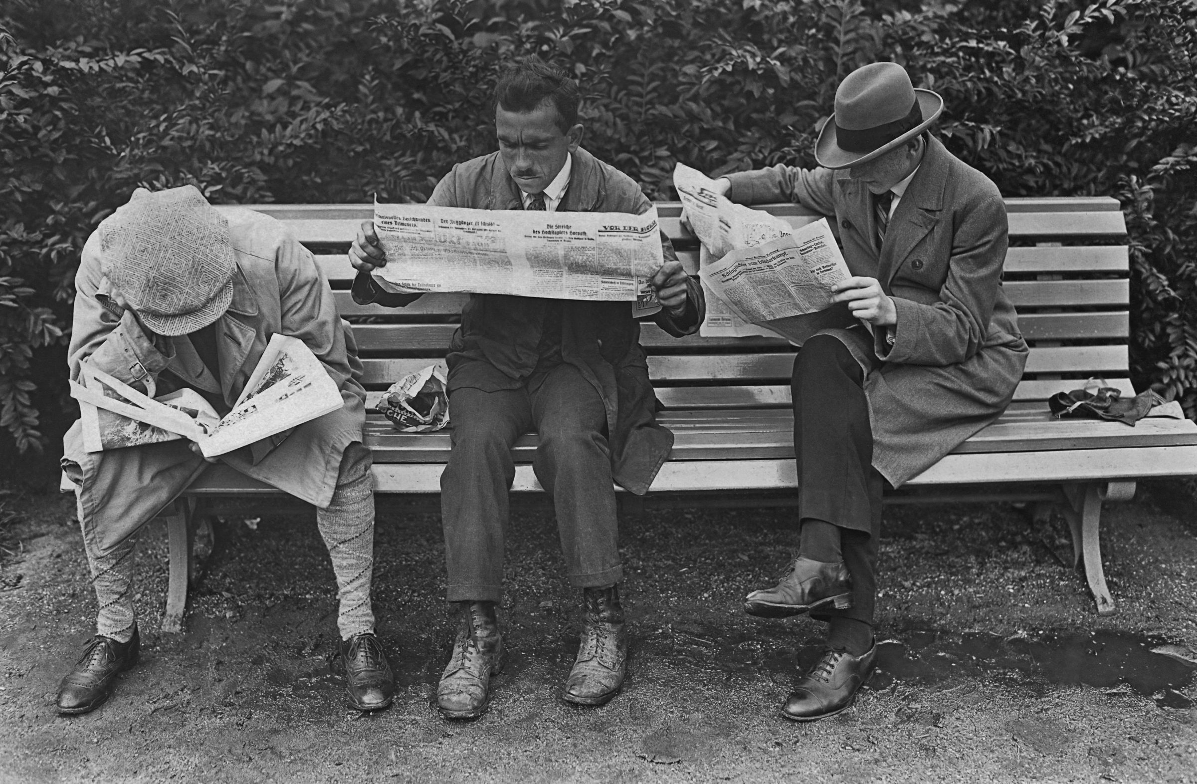Three men sitting on a park bench reading the newspaper