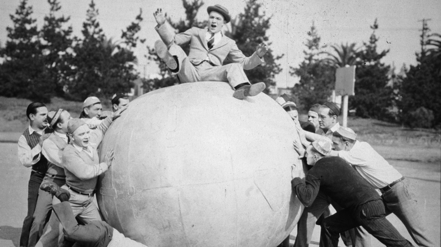 Two groups of men push on opposite sides of a large ball, on top of which sits a man who is losing his balance in a still from the silent film, 'Going Great'. (Photo by Hulton Archive/Getty Images)