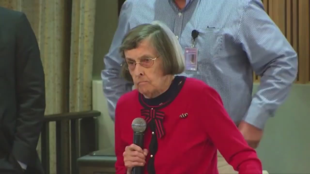 A photo of Greta Rogers as she spoke at a Phoenix City Council meeting in 2018. She is wearing a red sweater and holding a microphone. She also is wearing glasses. She has silver hair, cut short.