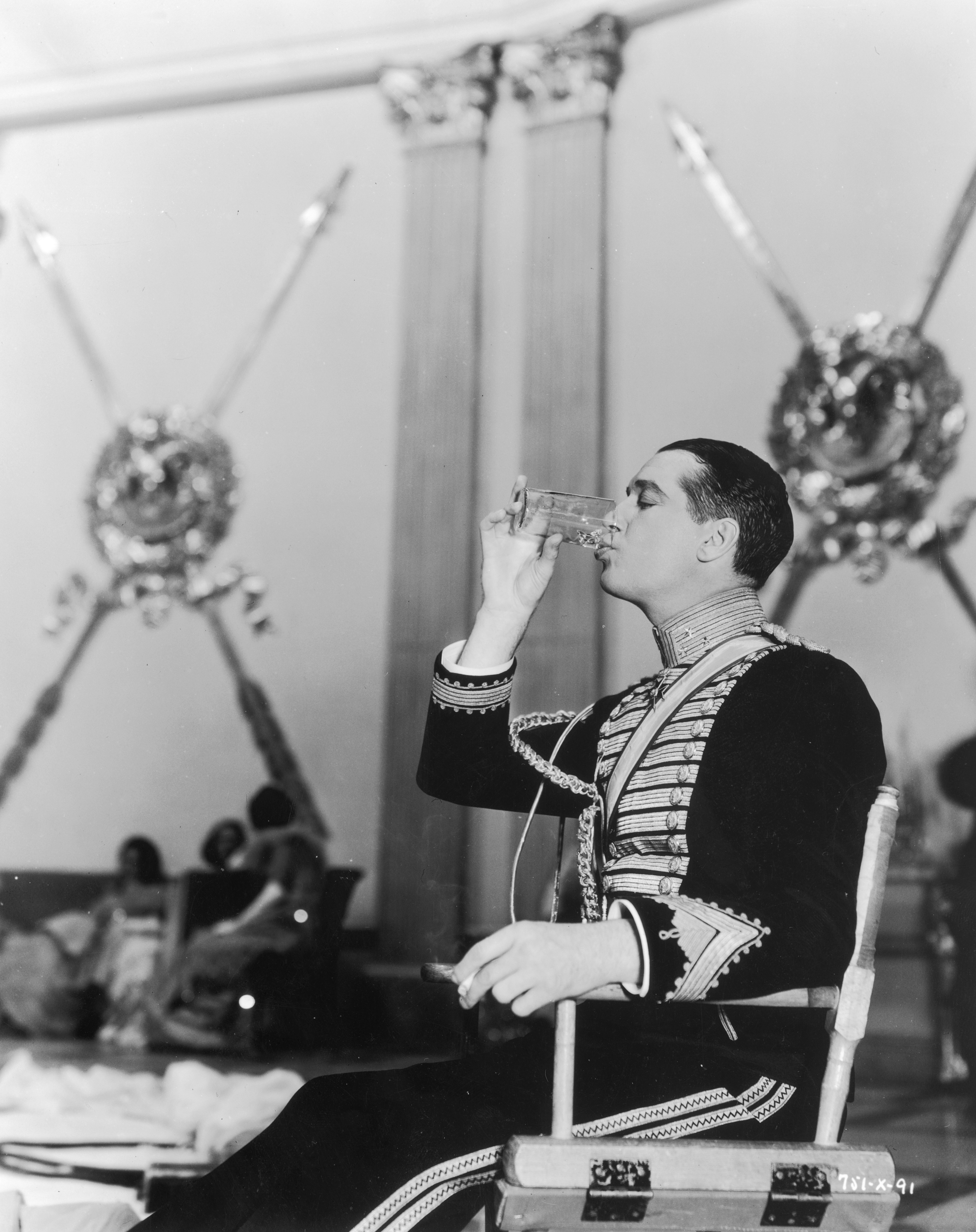 1934: Profile view of French actor Maurice Chevalier drinking a glass of water while seated in a folding chair on the set of director Ernst Lubitsch's film, 'The Merry Widow'. Chevalier wears a military uniform.