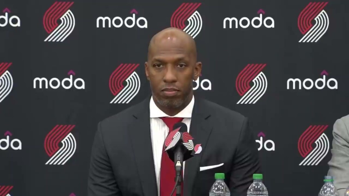 A photo of new Portland Trail Blazers head coach Chauncey Billups. He is wearing a suit and tie in Blazers colors. Behind him is a backdrop with the Blazers logo.