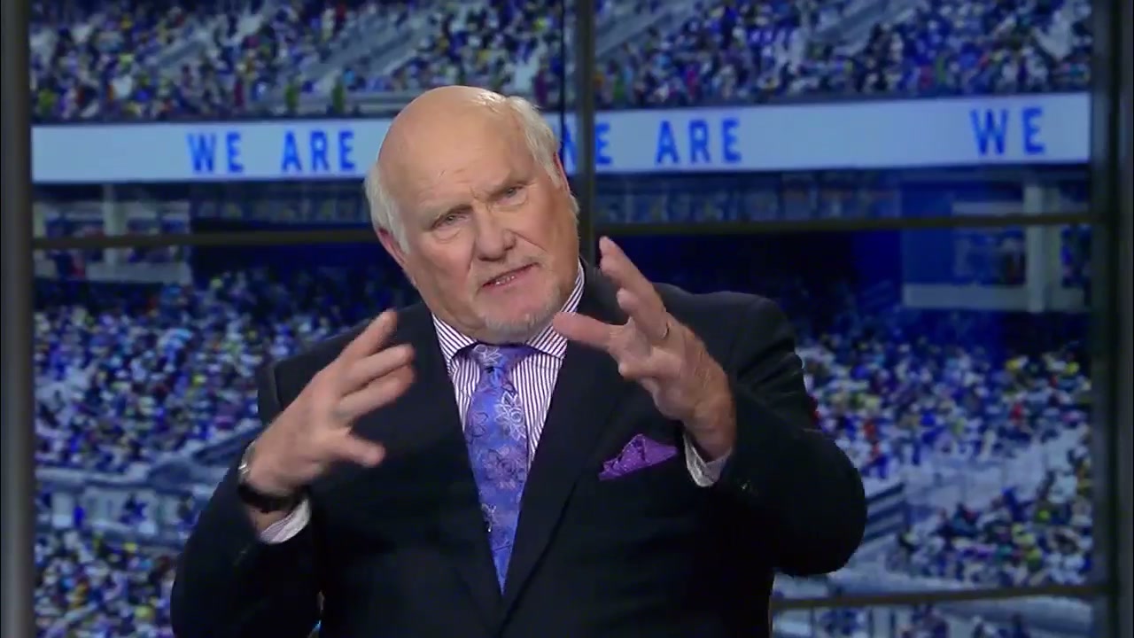 Terry Bradshaw, in a black suit and purple tie, holding his hands up like he's holding a pumpkin (there's nothing in his hands)