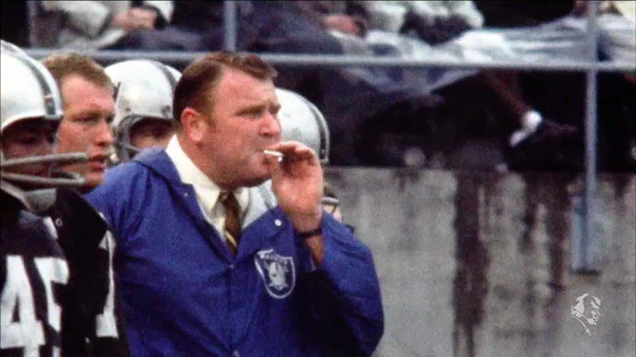 John Madden in shirt and tie and vinyl Raiders jacket, smokes on the sidelines
