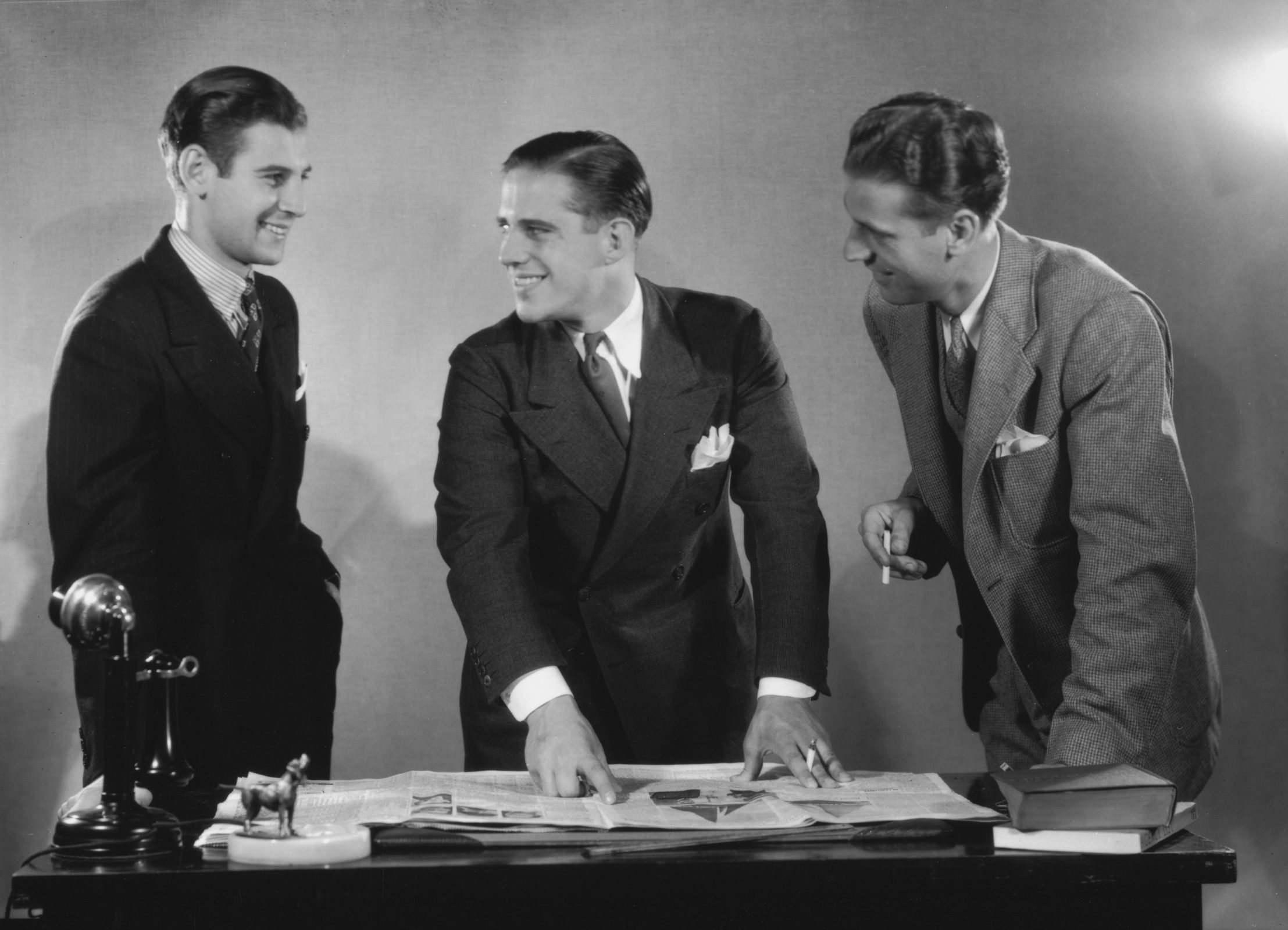 Three men in suits smiling as the man in the centre points to an article in a newspaper, which is spread out on the desk before them, circa 1945. Also on the desk are a telephone, an ashtray, and a small pile of books. (Photo by Archive Photos/Hulton Archive/Getty Images)