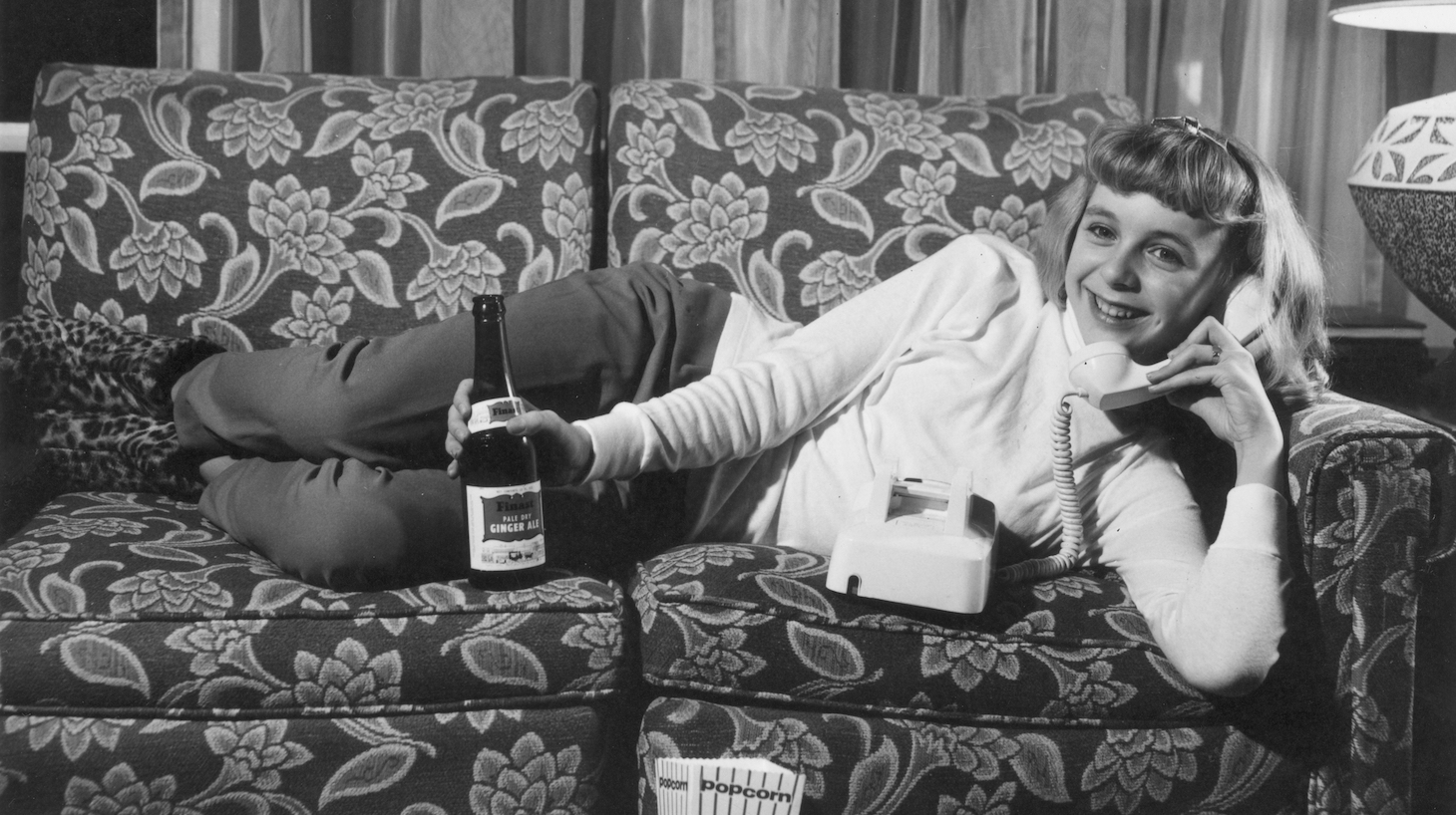 circa 1955: Portrait of a teenage girl lying on a sofa with a bottle of ginger ale and a telephone. A box of popcorn sits in front of her.