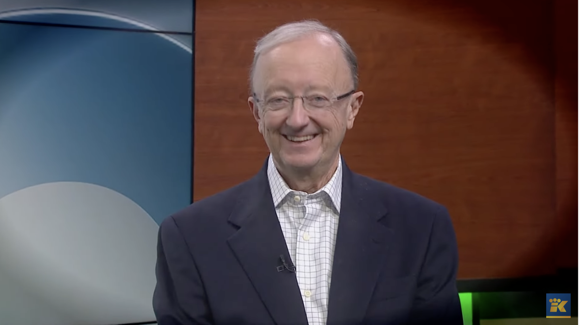 A photo of John Clayton. He is sitting in a TV studio, wearing a suit, and smiling.