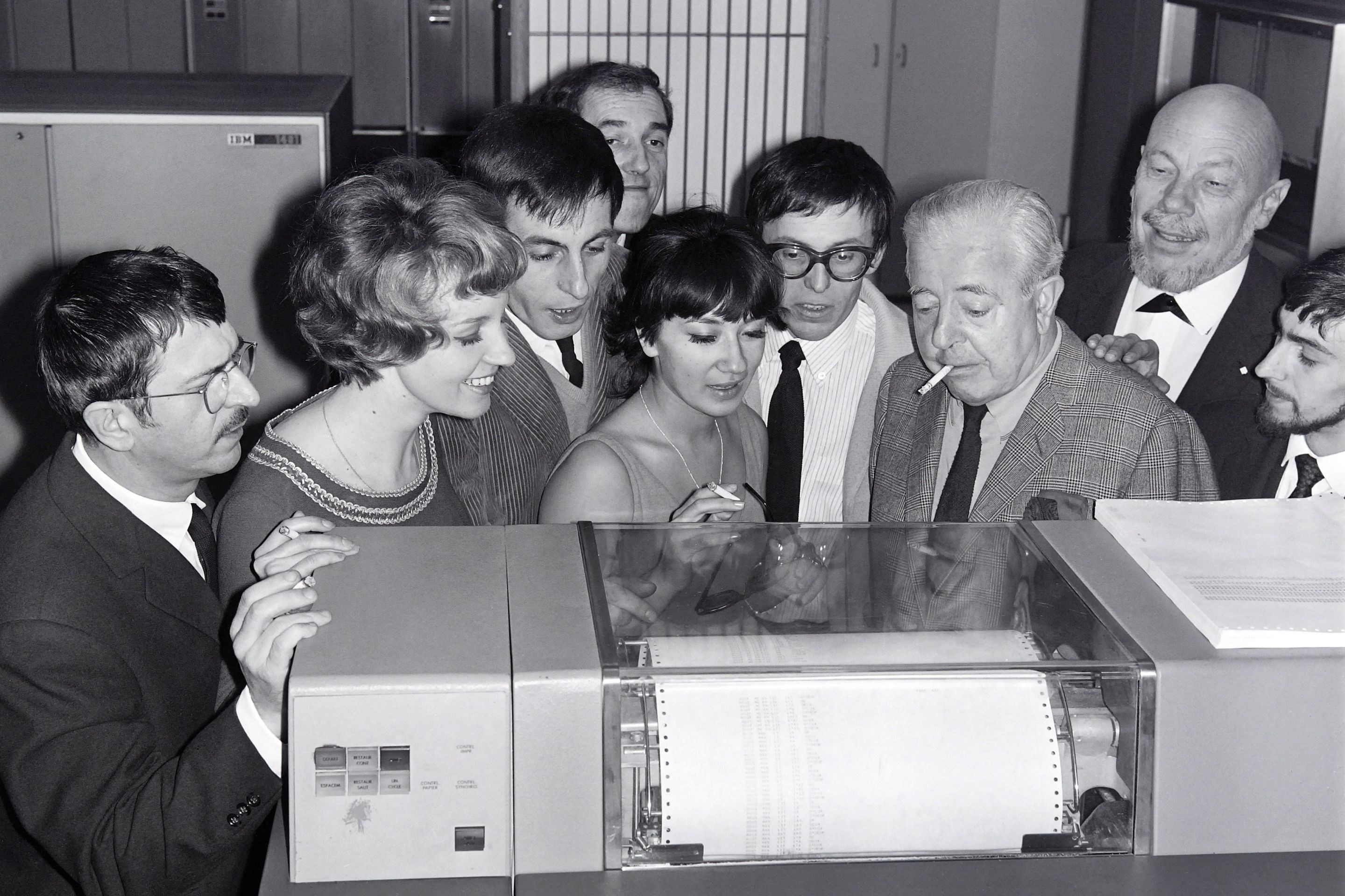 (From L to R) Roger Riffard, Lise Granvel, Armand Babel, Jean-Pierre Dutour, Catherine Sotha, Romain Bouteille, Leo Campion, Jacques Prevert and Jacques Barbier look at the IBM computer which will find the title of the new play written by Romain Bouteille (4thR with glasses), on November 13, 1965 in Paris. (Photo by - / AFP) (