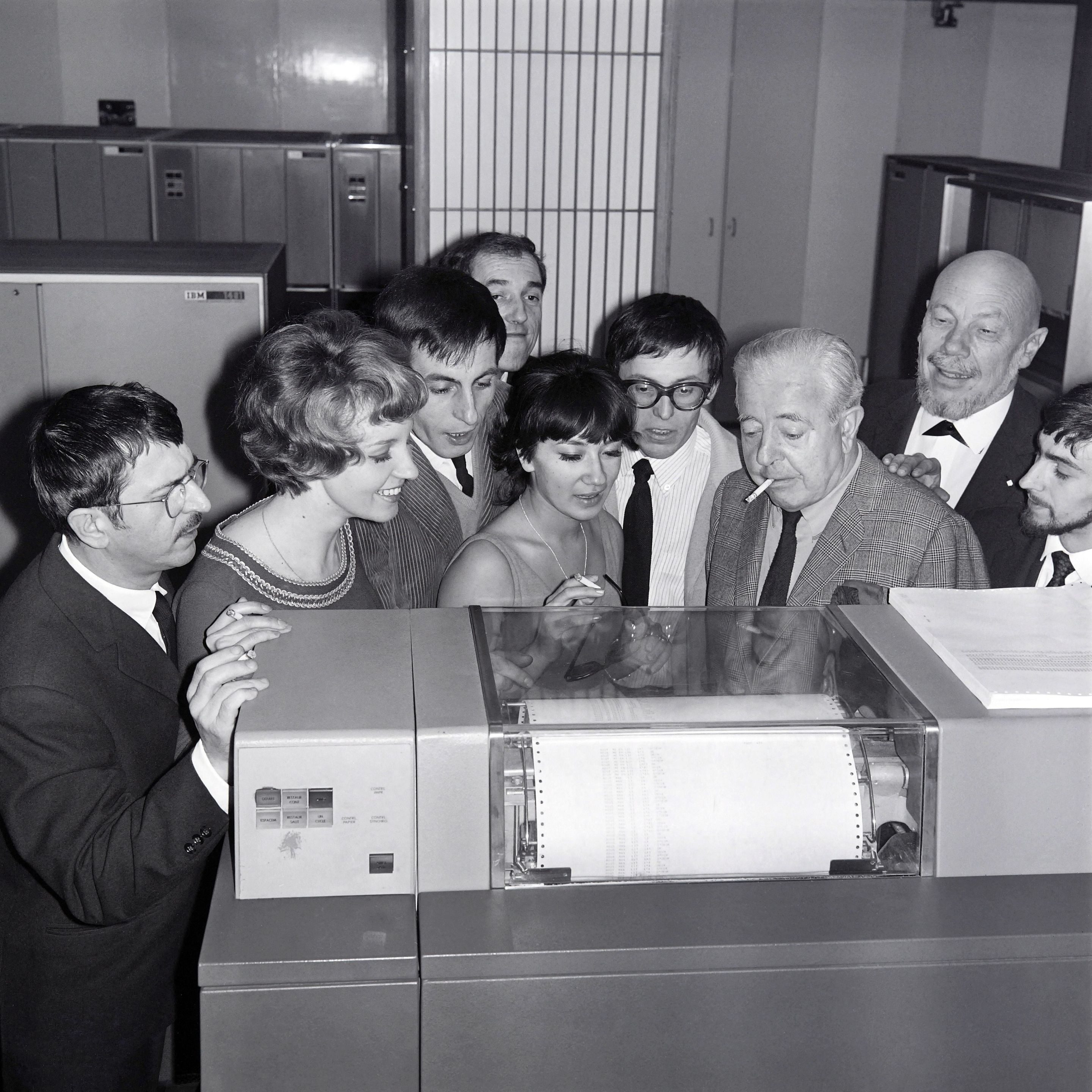 (From L to R) Roger Riffard, Lise Granvel, Armand Babel, Jean-Pierre Dutour, Catherine Sotha, Romain Bouteille, Leo Campion, Jacques Prevert and Jacques Barbier look at the IBM computer which will find the title of the new play written by Romain Bouteille (4thR with glasses), on November 13, 1965 in Paris. (Photo by - / AFP) (