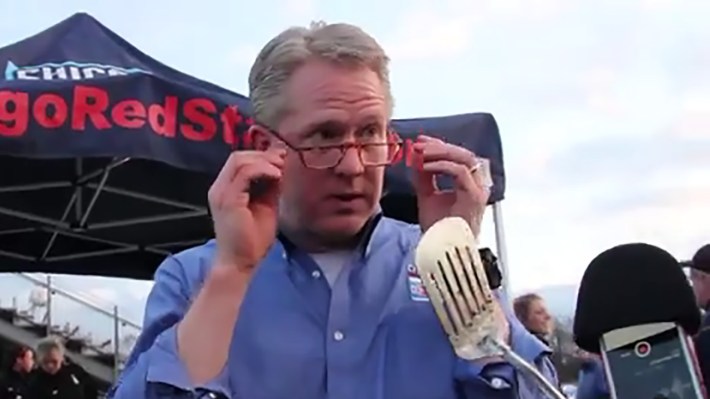 A screencap of Arnim Whisler hanging out before a Chicago Red Stars game. The image is from a promo video made by the Red Stars and was pretty silly, which is why he is speaking in front of a microphone and a spatula. Otherwise, he is an older white man with graying hair. In the image, you can see he is wearing red glasses and a blue button-down T-shirt.