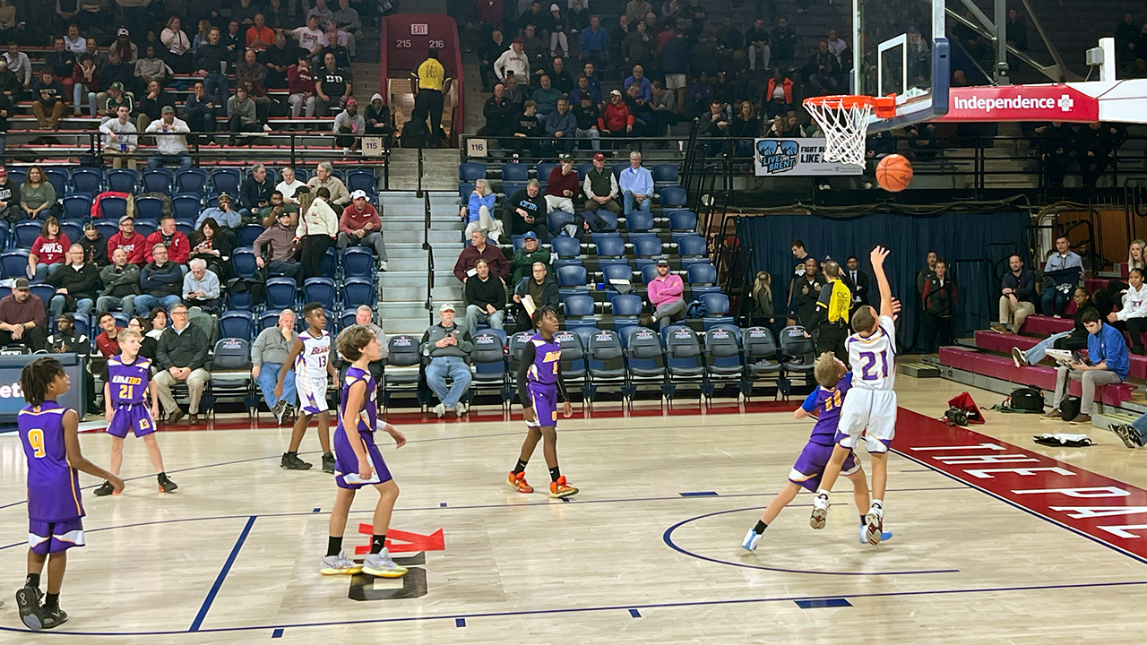 Little kids play basketball at The Palestra. Light brown court, red baseline. Purple and white teams, white team kid shooting against a purple team kid, it's a layup