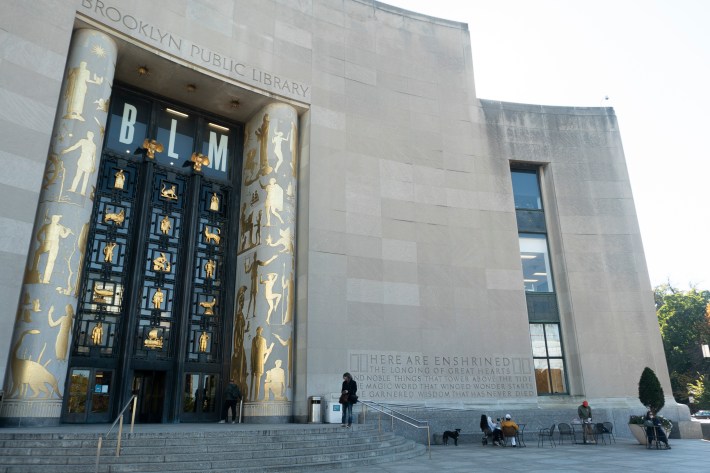 The exterior of the Brooklyn Public Library. It has large doors and pillars with figures of human beings and animals on them in gold. Near the top of the doors are the letters BLM. A ew people are sitting on the steps out front.