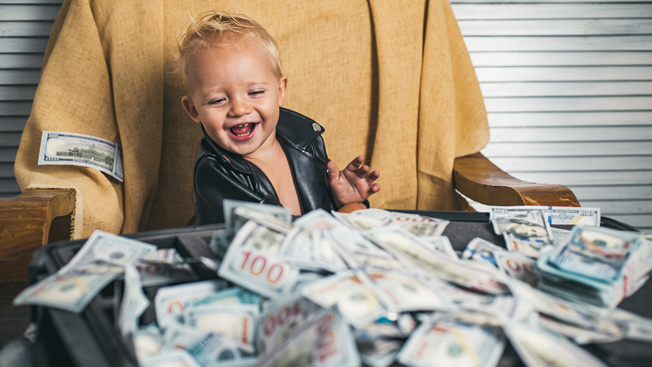 A baby, blonde hair, pretty cute, but not like incredibly cute, sititng on a chair with a tan blanket draped over it. the baby is wearing a leather jacket. there's a bunch of money in front of him. it all kinda looks real except for one $100 bill which looks so fake so pretend that bill looks real too