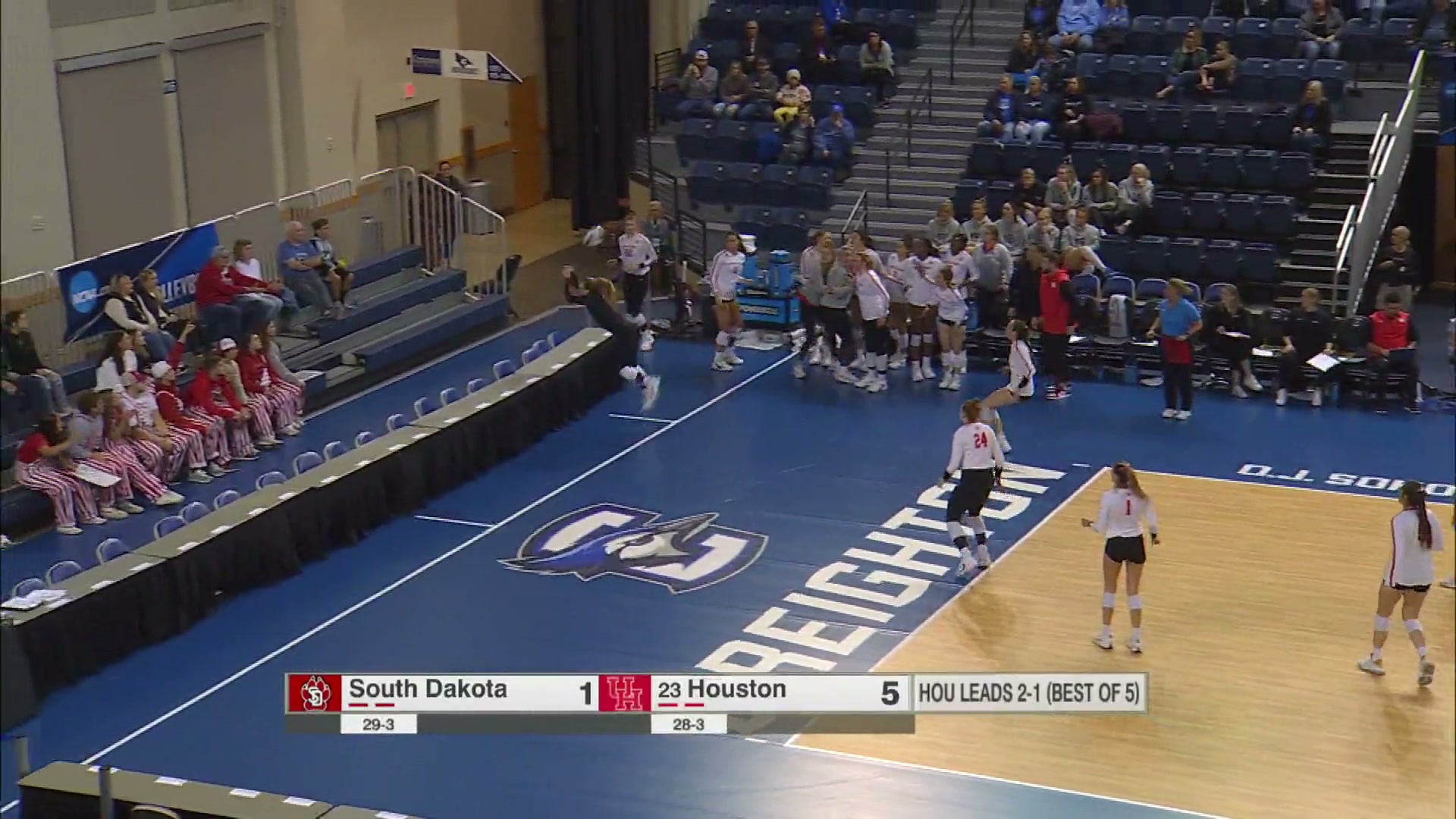 Volleyball player dives into table. Blue court, table is press row but empty. South Dakota 1 point vs Houston 5 points Houston leads 1-2 (Best of 5)