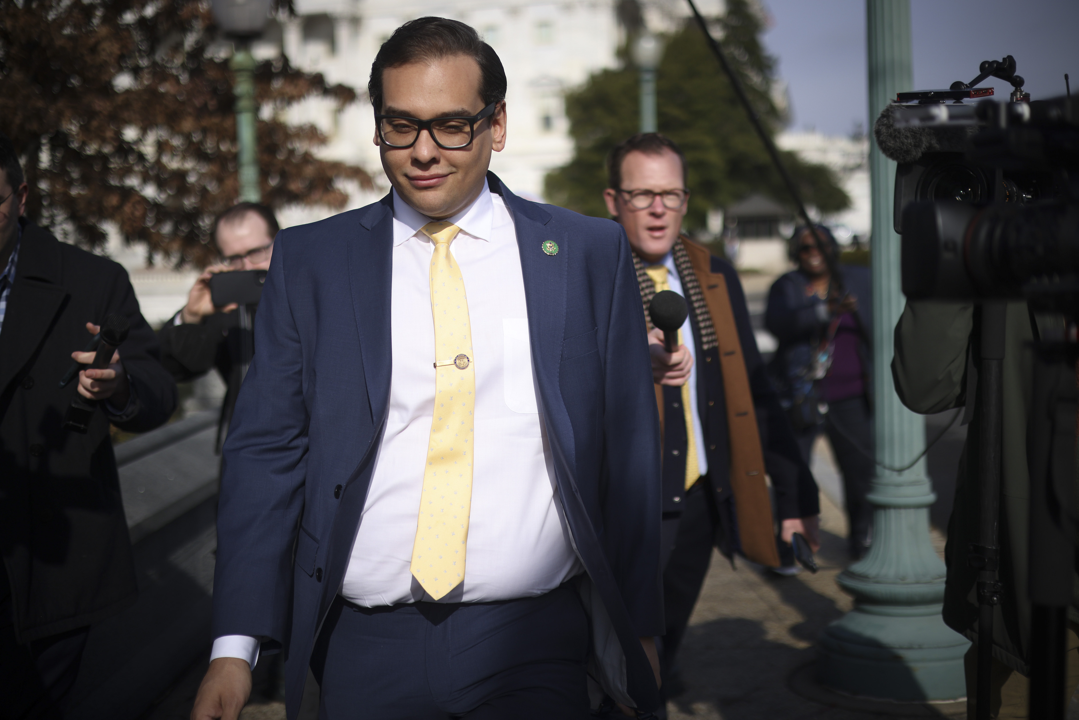 Pre-disgraced Rep. George Santos walking around near the House of Representatives making a silly face while media people say "what about your lies, sir."