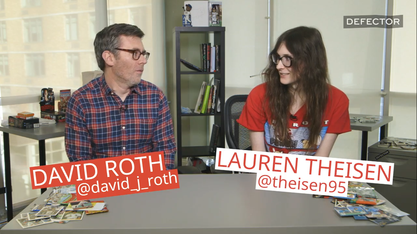 A screenshot of David Roth and Lauren Thiesen as they sit at a table to open up a pack of cards and remember some guys. Lauren is wearing a bright red graphic T-shirt. Roth, of course, is in flannel.