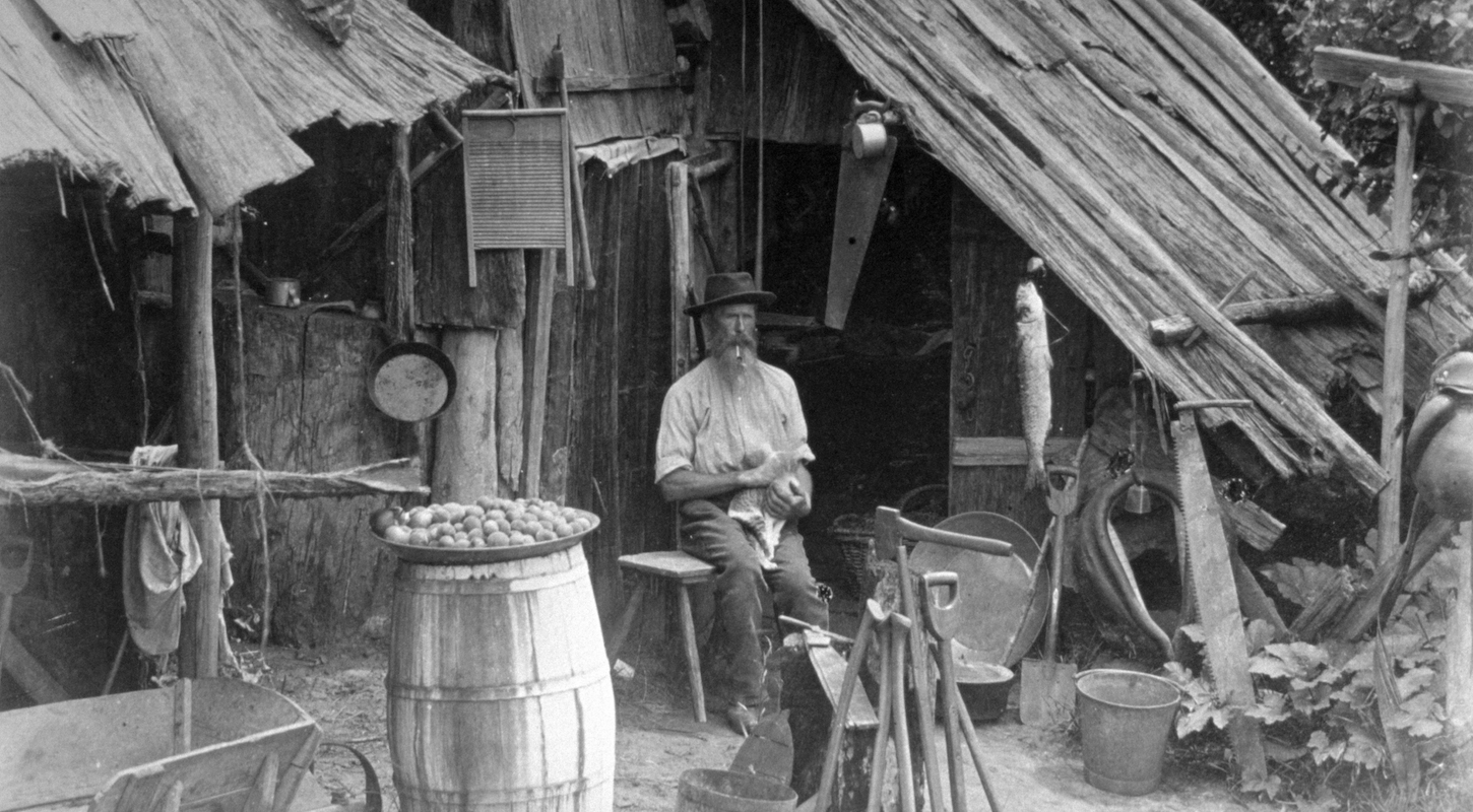 A grizzled prospector sits in front of a hovel in an old black-and-white photo