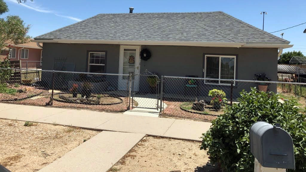 A single-story ranch house surrounded by a chain link fence. It is pleasant and well-groomed. There is a mailbox in the front of the photo.