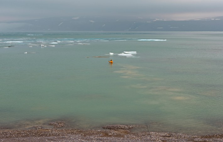 A photo of a pale green and blue ocean, with a tiny human figure dressed in orange setting up a hydrophone while submerged.