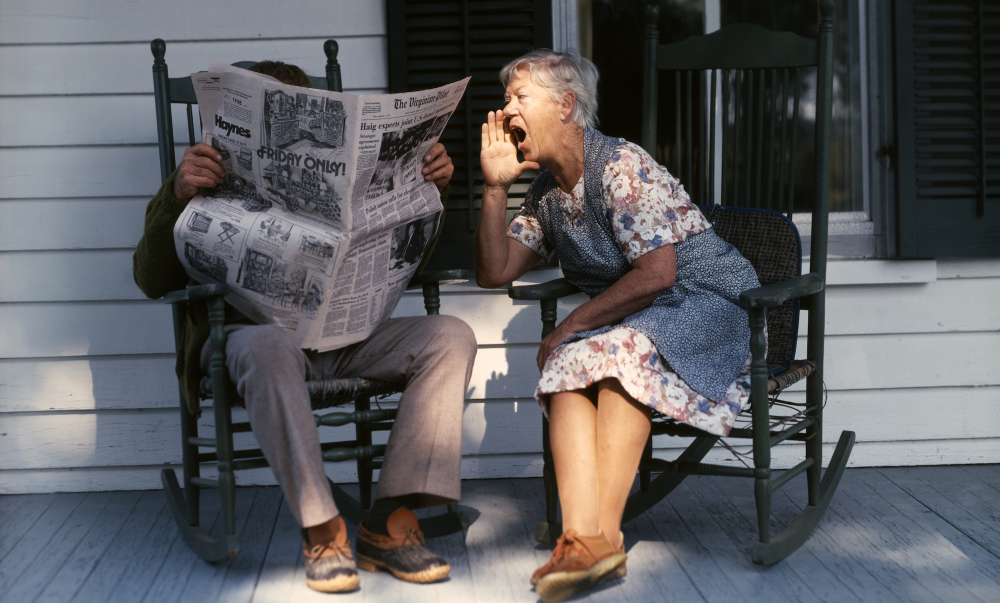 1970s SENIOR COUPLE SITTING ON PORCH ROCKING CHAIRS MAN READING NEWSPAPER WOMAN SHOUTING AT HIM (Photo by D. Corson/ClassicStock/Getty Images)
