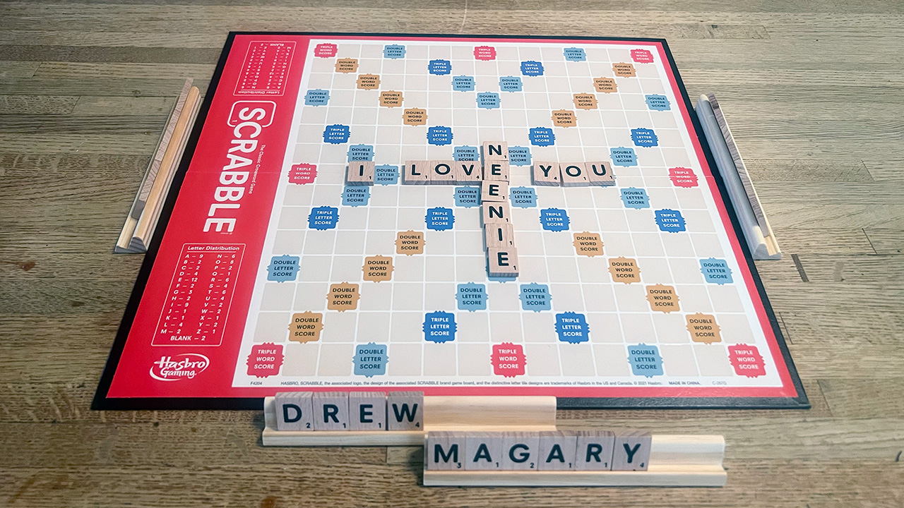 A Scrabble board with "I LOVE YOU" across and "NEENIE" down. In front are two tile racks with "DREW MAGARY" on them.