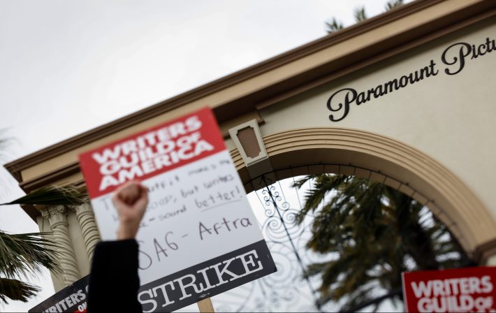 As seen through a tilt-shift lens, supporters of the Writer's Guild of America strike, picket along Melrose Avenue, at Paramount Studios, in Los Angeles, CA, Tuesday, May 9, 2023. In the background the sign says Paramount. In the front is a raised fist.