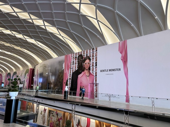 A kinda empty mall, though there are two people walking in it. A big sign promises: GENTLE MONSTER: COMING SOON.