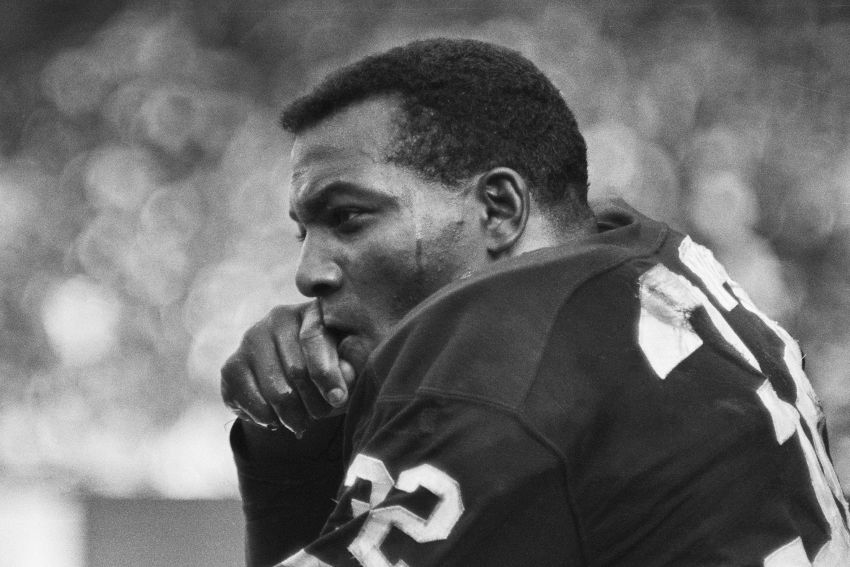 A black and white photo of a young Jim Brown, when he played for the Cleveland Browns. It shows Brown sitting on a bench, with fans off in the blurry distance.