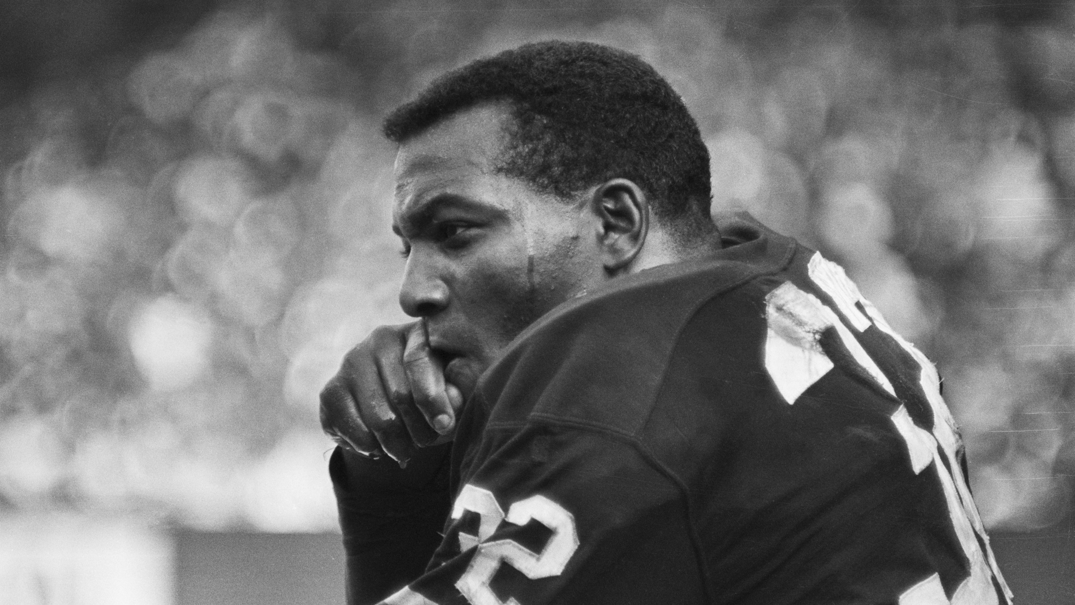 A black and white photo of a young Jim Brown, when he played for the Cleveland Browns. It shows Brown sitting on a bench, with fans off in the blurry distance.