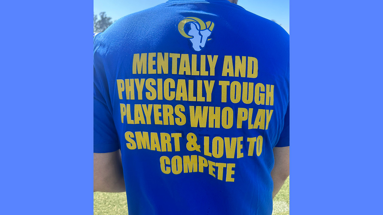 Back of a man wearing a shirt with the Rams logo on it and it says "MENTALLY AND PHYSICALLY TOUGH PLAYERS WHO PLAY SMART & LOVE TO COMPETE"