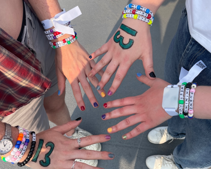 Two pairs of hands, each with a '13' drawn on them, multiple friendship bracelets, light-up bracelets, and fingernails painted in correspondence to Taylor Swift's ten eras.