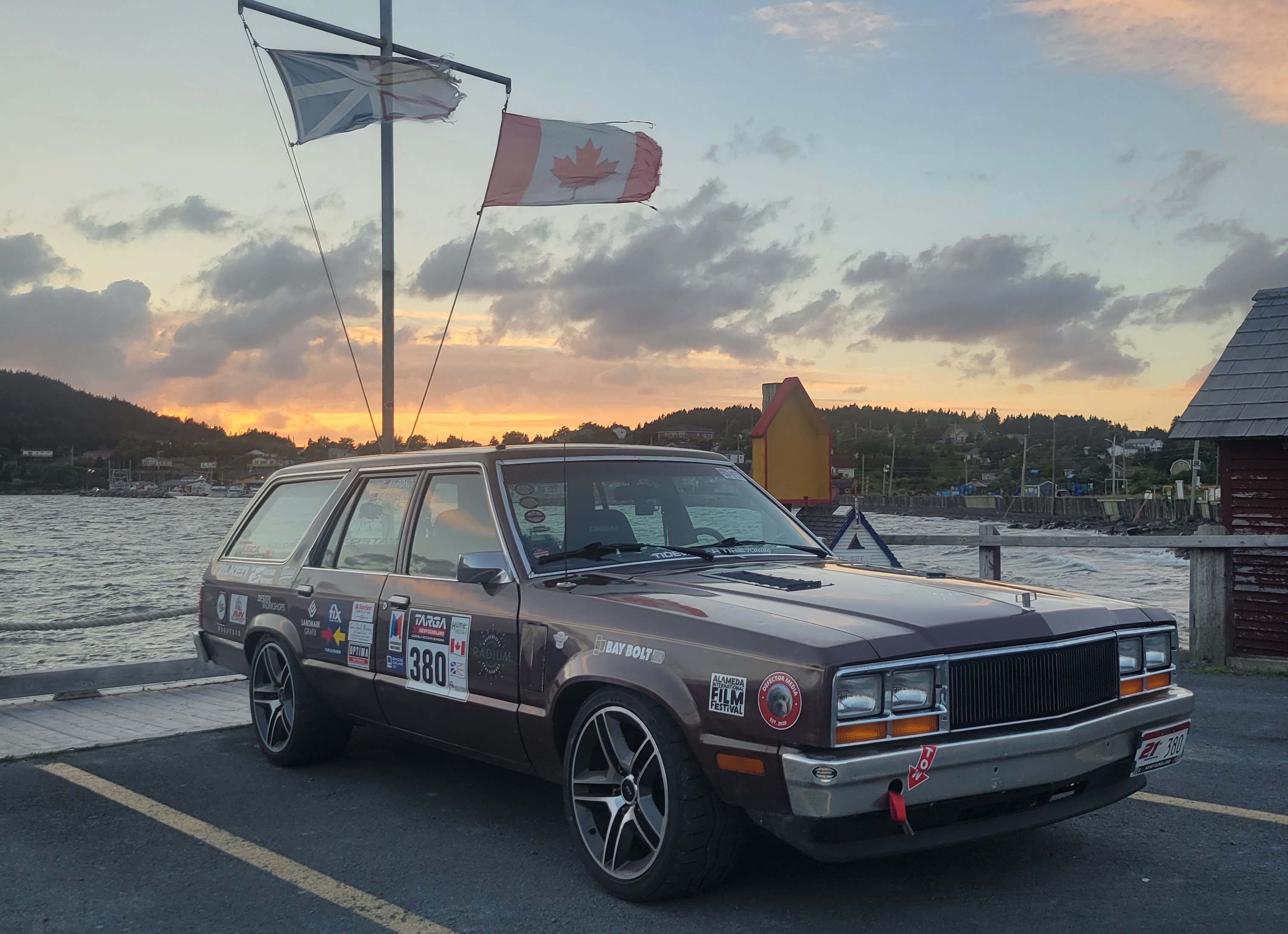 A photo of a brown stationwagon next to the water. It is covered in stickers.