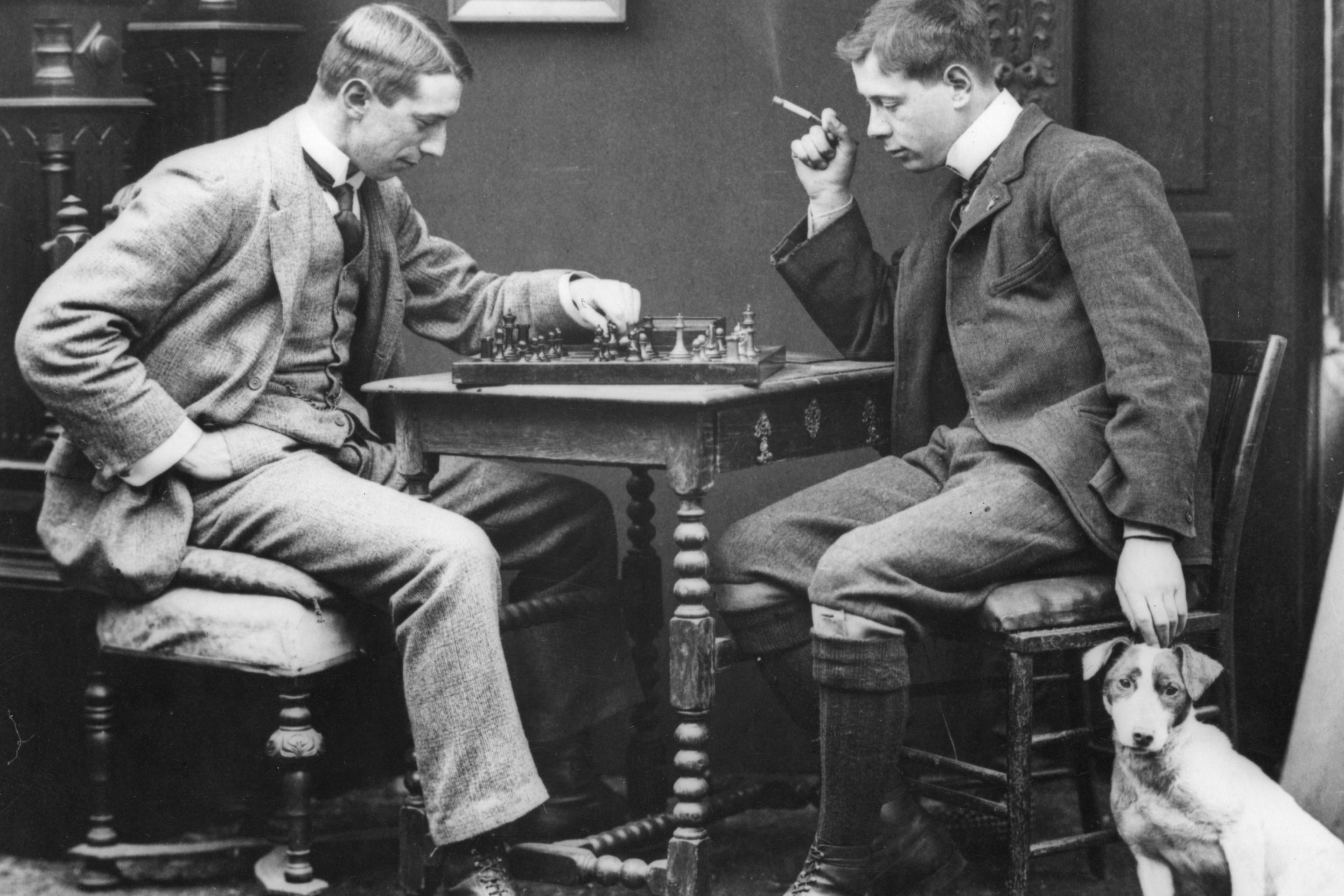circa 1885: A couple of men playing a leisurely game of chess. The photo is in black in white. The men are dressed in suits. One of them is also petting a dog.