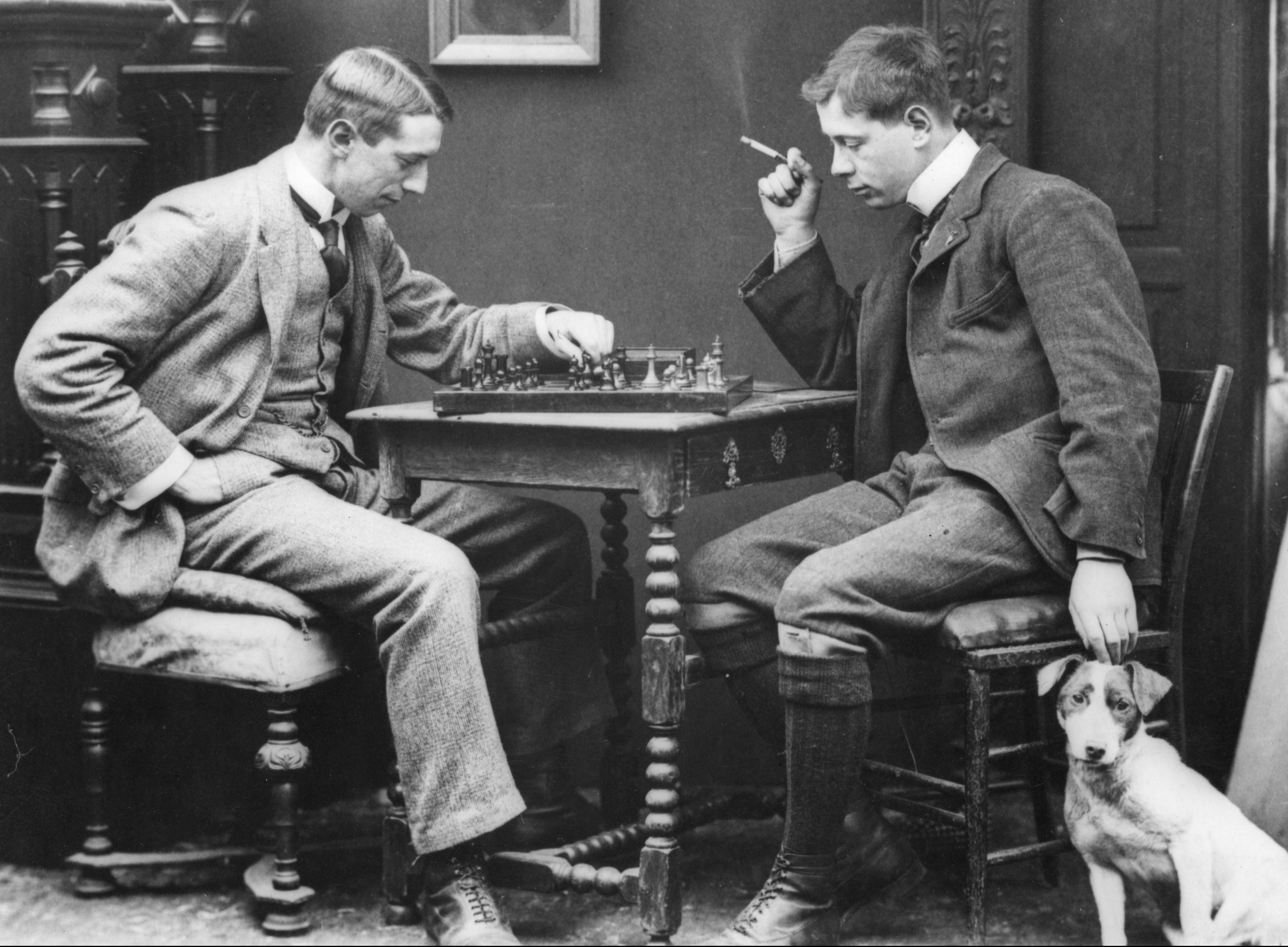 circa 1885: A couple of men playing a leisurely game of chess. The photo is in black in white. The men are dressed in suits. One of them is also petting a dog.