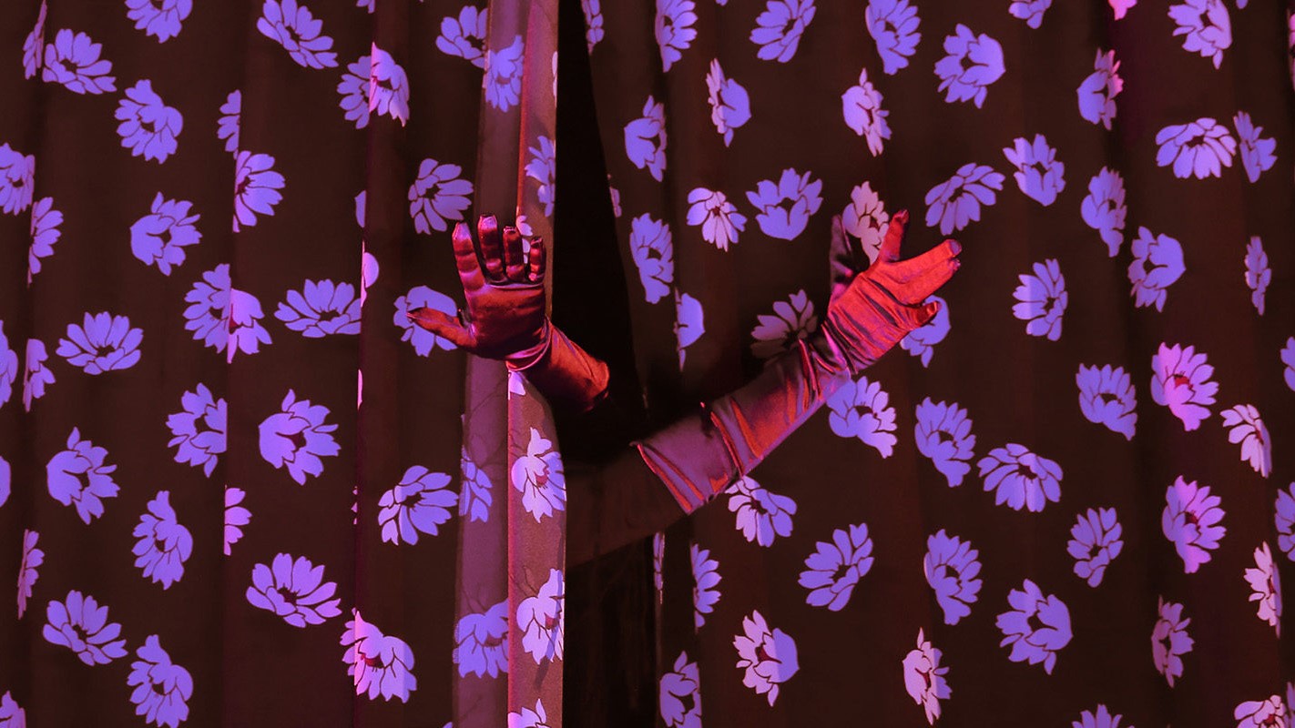 gloved hands reaching out from behind a floral curtain