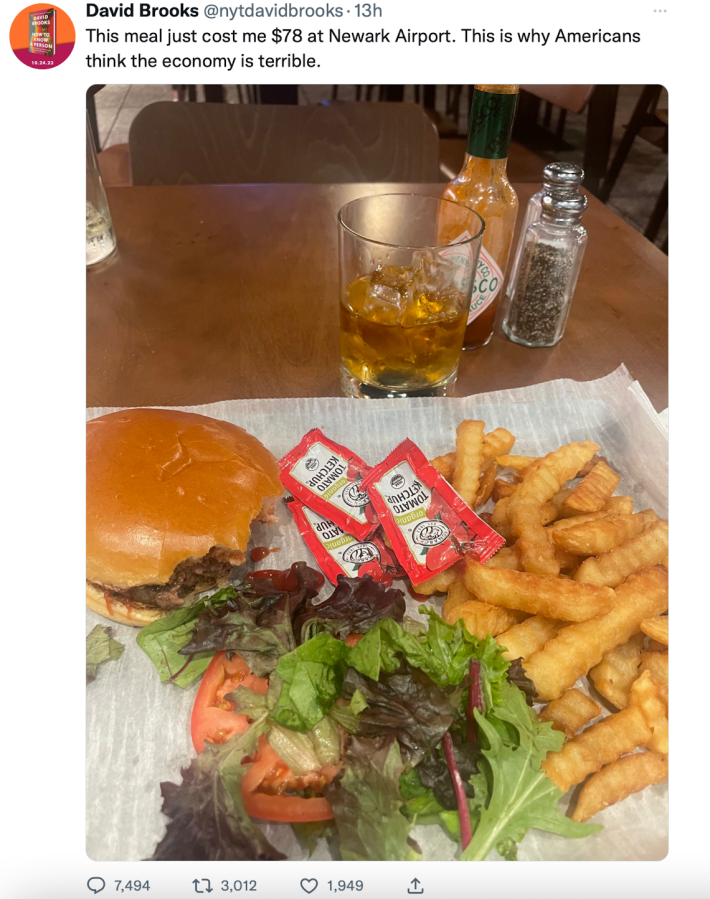 A tweet from NYT's David Brooks. A photograph shows a burger, french fries, a salad, and what appears to be a glass with whiskey on the rocks in it; the text says "This meal just cost me $78 at Newark Airport. This is why Americans think the economy is terrible."