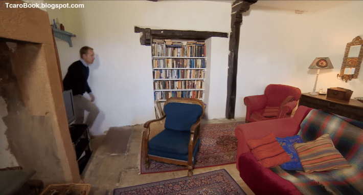 A very ugly room with bare white walls, but featuring a charming recessed bookcase in the middle of the frame. A man is entering the frame on the left, where you can just barely see, poking out from around the side of what appears to be a huge plaster hearth (?), the edge of a comically small flat-panel TV that would be visible to a likewise comically small portion of the room.