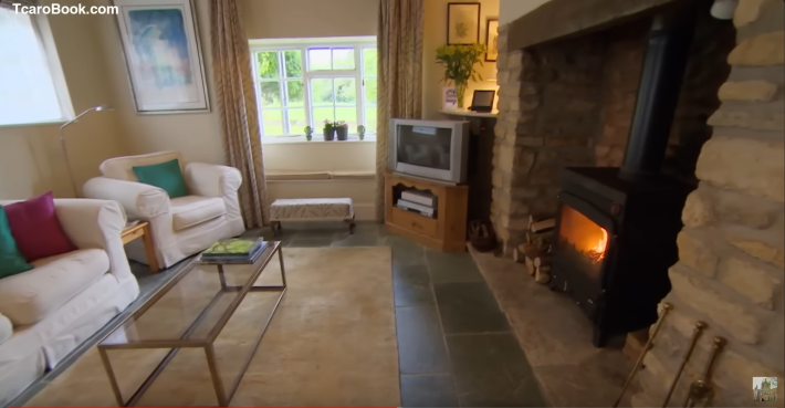A room featuring an absolutely gigantic stone hearth, taller than a normal adult, hosting a wood-burning stove. Against the far wall, on a hideous little wood-paneled end table thing, sits a gray cathode-ray tube TV of the kind that was newish circa the year 2002.