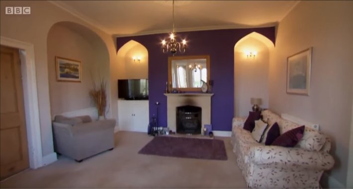 A very weird and ugly British living room with a hideous purple accent wall, in which a small TV sits in a recessed archway in a corner, facing an armchair, all but totally invisible to the sofa that provides 75 percent of the room's seating.