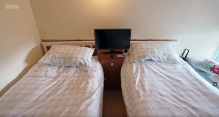 A bleak under-decorated British bedroom with two parallel single beds facing the same direction. In between them, on a nightstand, is a small flatscreen TV... facing the same direction as the beds, so that someone laying with their head on the pillow on either bed could not hope to see the screen, which would be by their ear.