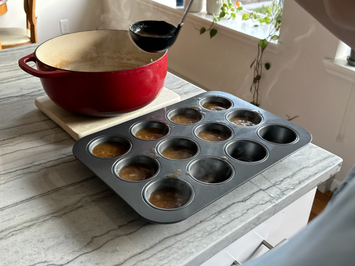 Ladling soup from a pretty red Dutch oven into cupcake tins on a granite countertop