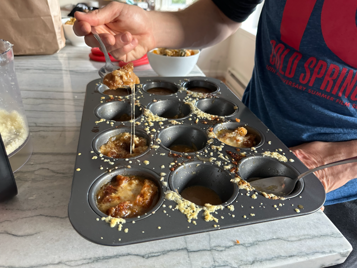 A spoonful of French onion soup being lifted out of a cupcake tin, trailing behind it strands of stretchy gruyere cheese.