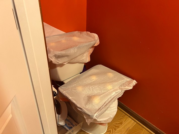 Two trays of raw dough balls, covered in white kitchen trash bags, rest on a toilet in a small bathroom.
