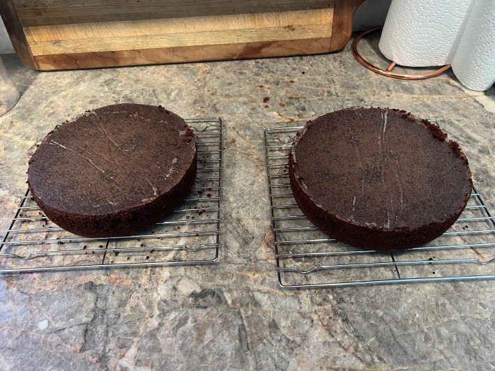 Two very dark chocolate sponges, lined on the bottom with parchment paper, resting on cooling racks.